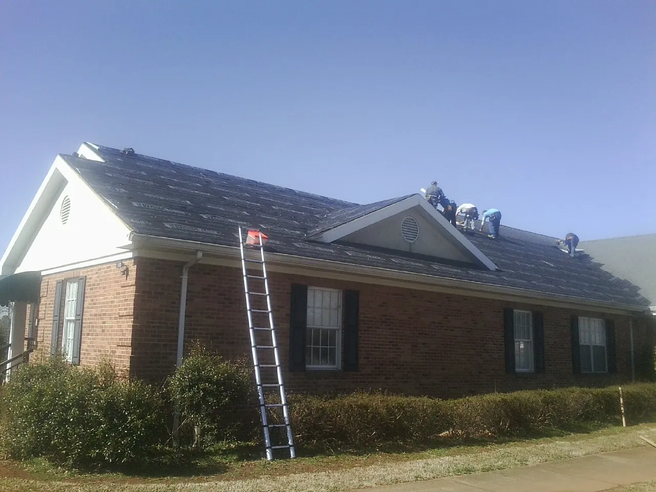 Crew tearing shingles off a brick ranch roof with a ladder set — Emergency Roof Repair work by our crew in Rush Springs
