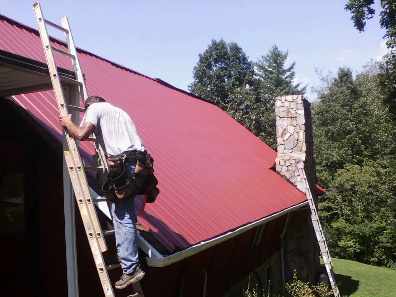 Roofer on a ladder beside a red metal roof and stone chimney — Soffit & Fascia Repair by Roofing Brothers in Rush Springs